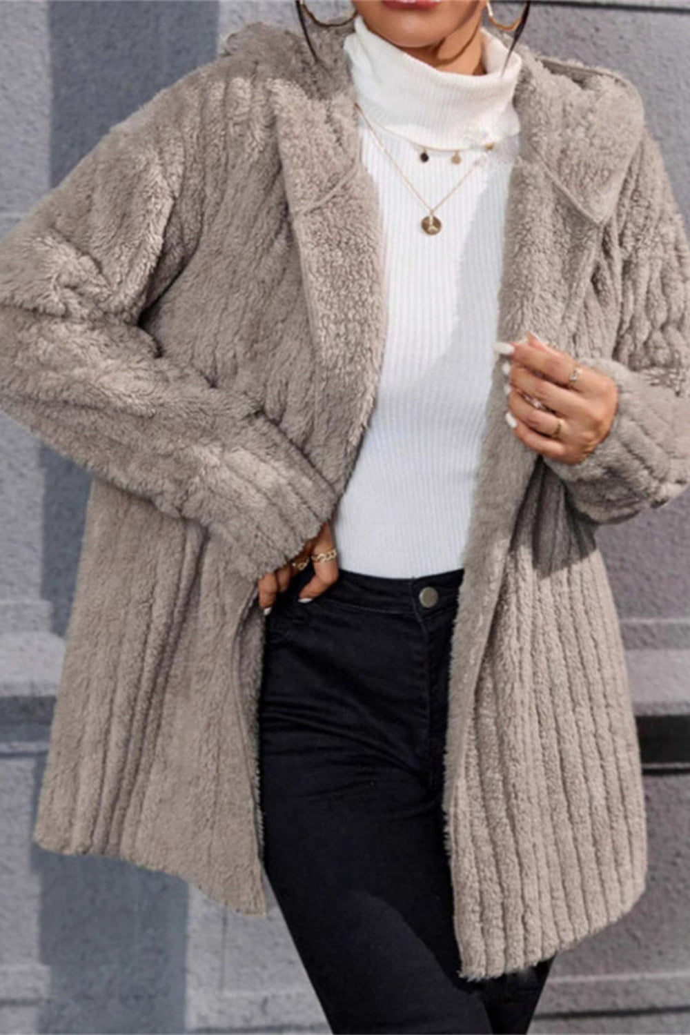 Person wearing a beige knitted cardigan over a white sweater and black pants against a gray background