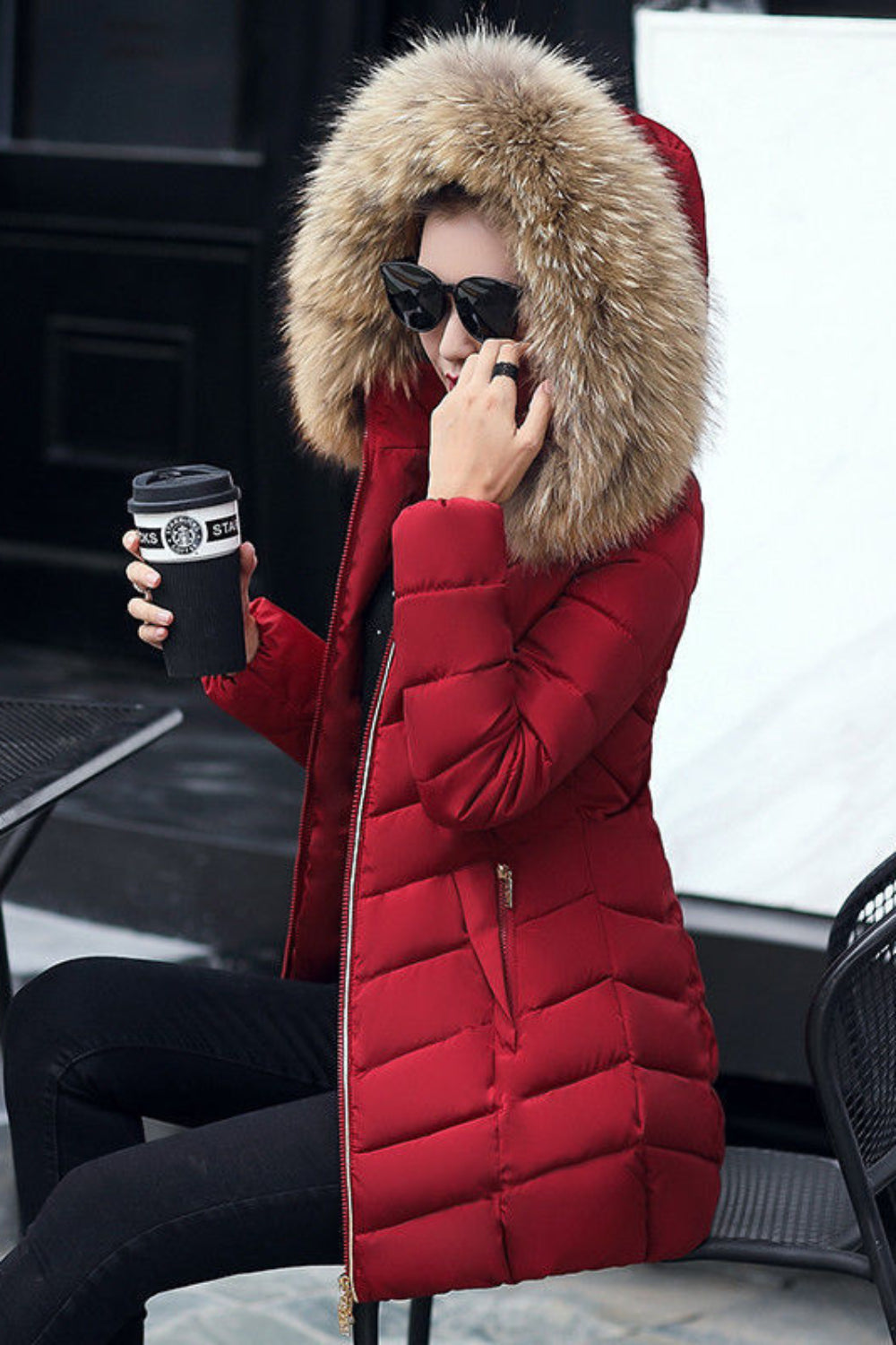 Person wearing a red puffer jacket with a fur hood, sitting outdoors holding a coffee cup.