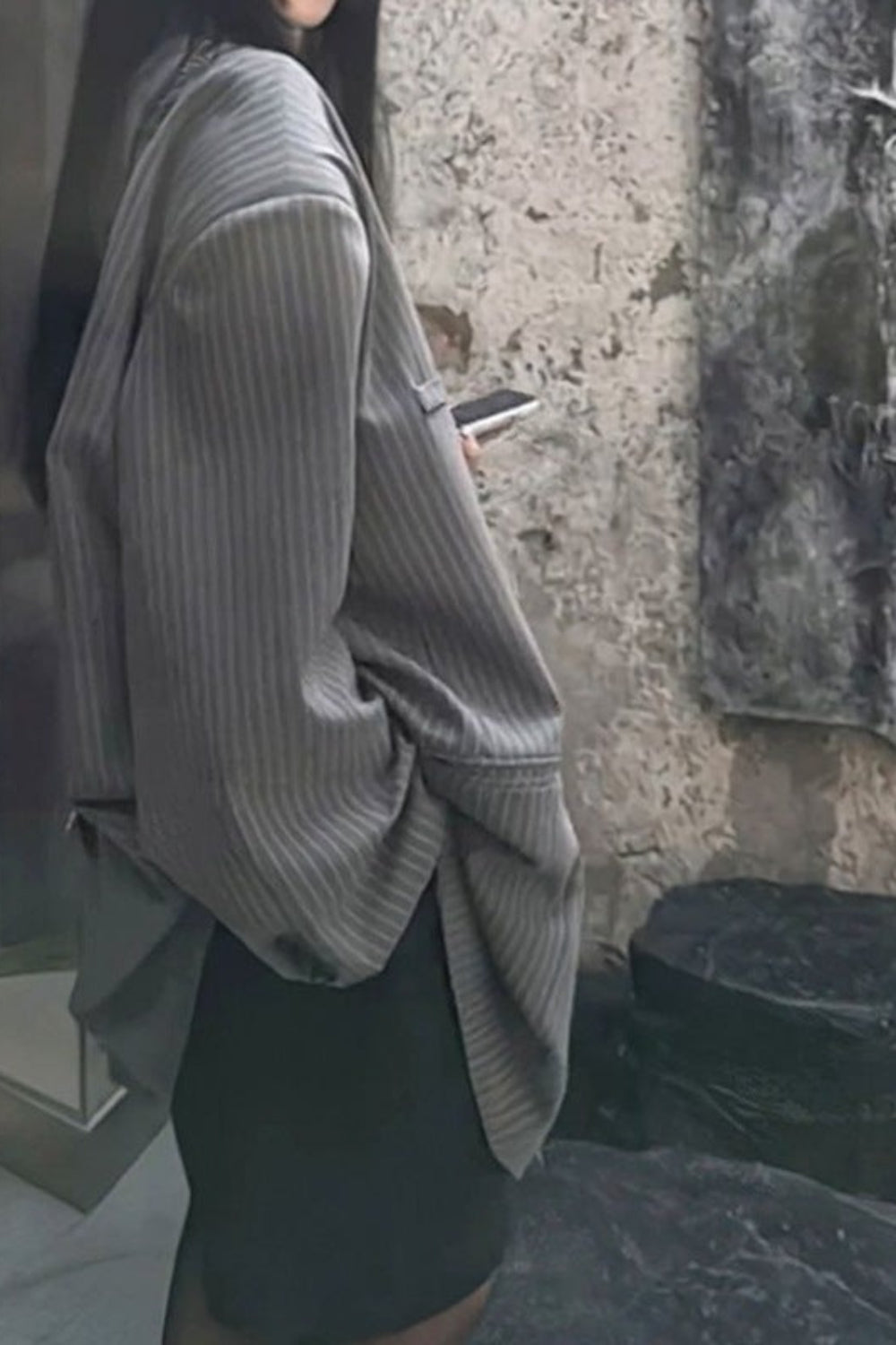 Person wearing a gray ribbed sweater and black skirt standing in front of a stone wall.