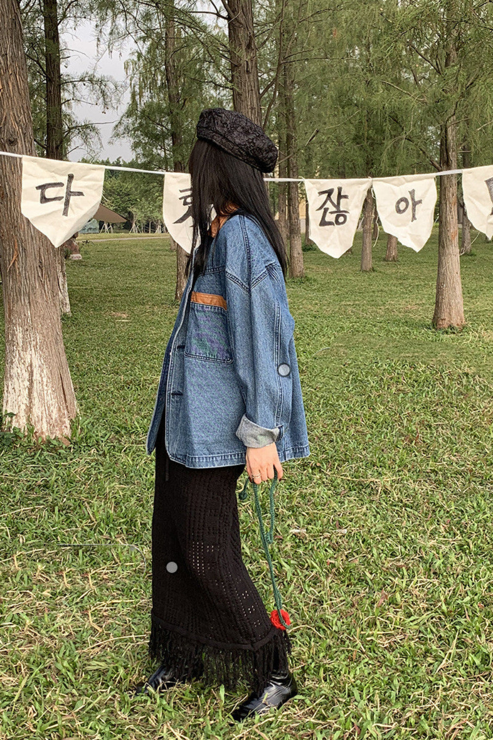 Person wearing a denim jacket and black skirt standing in a park with trees and banners in the background.