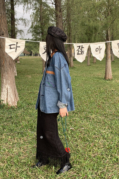 Person wearing a denim jacket and black skirt standing in a park with trees and banners in the background.