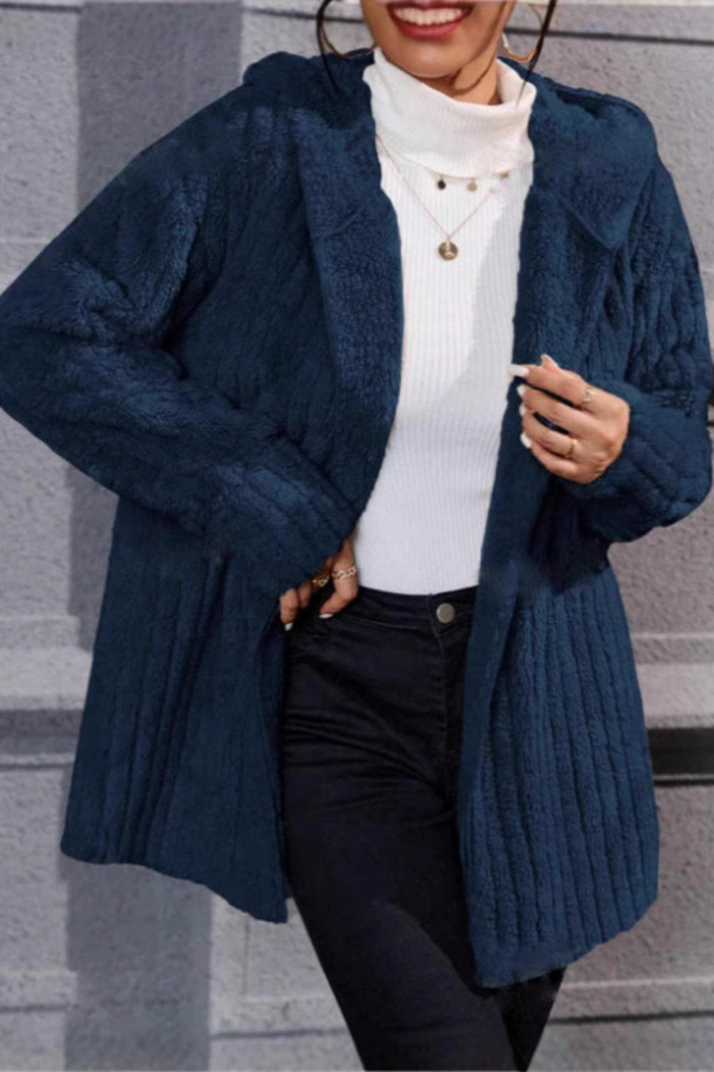 Person wearing a blue knitted cardigan over a white sweater and black pants, standing against a gray background.