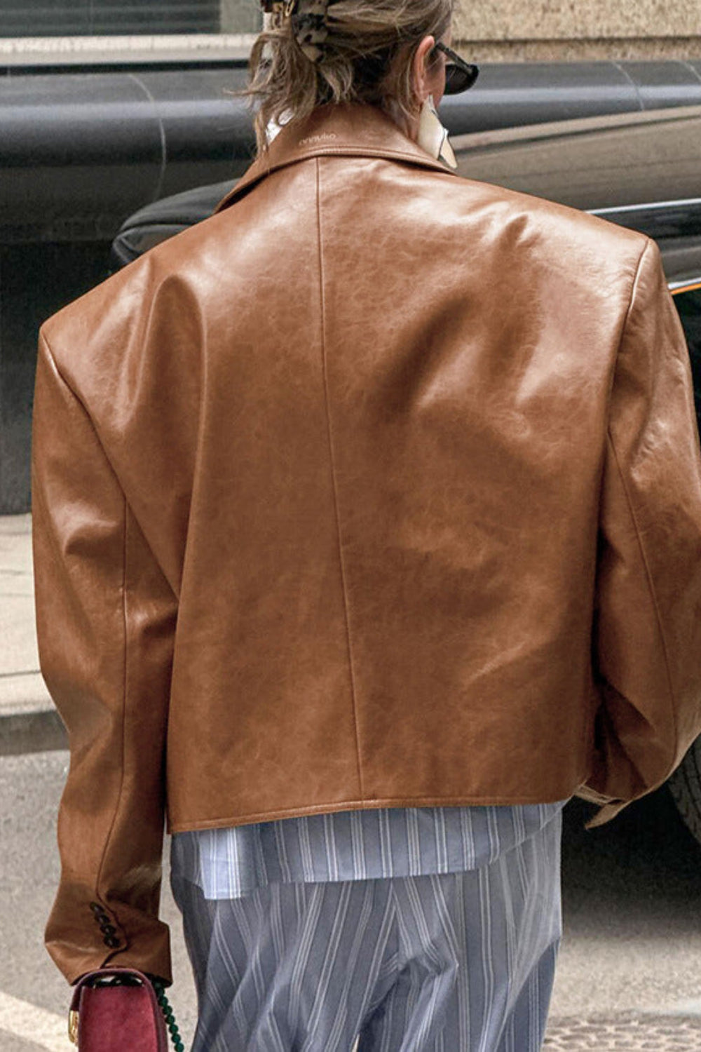 Person wearing a brown leather jacket walking on a street with a car in the background