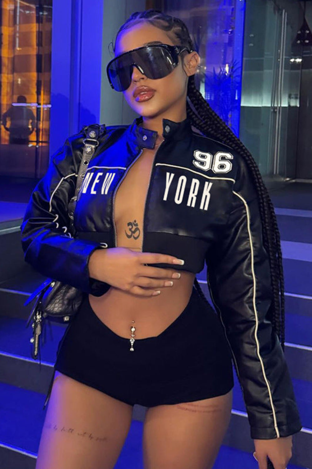 Person wearing a black jacket with 'New York' text, black shorts, and sunglasses in a blue-lit indoor setting.