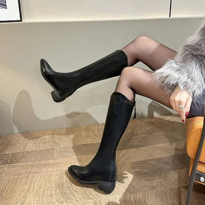 Person wearing black knee-high boots sitting on a chair with a neutral background