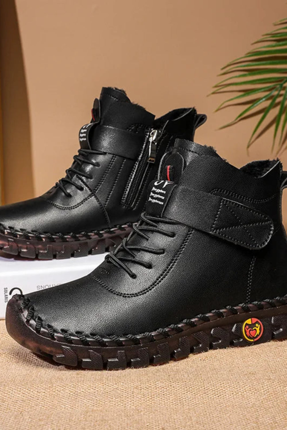 Black leather boots with a brand logo on a beige surface with a plant leaf in the corner.