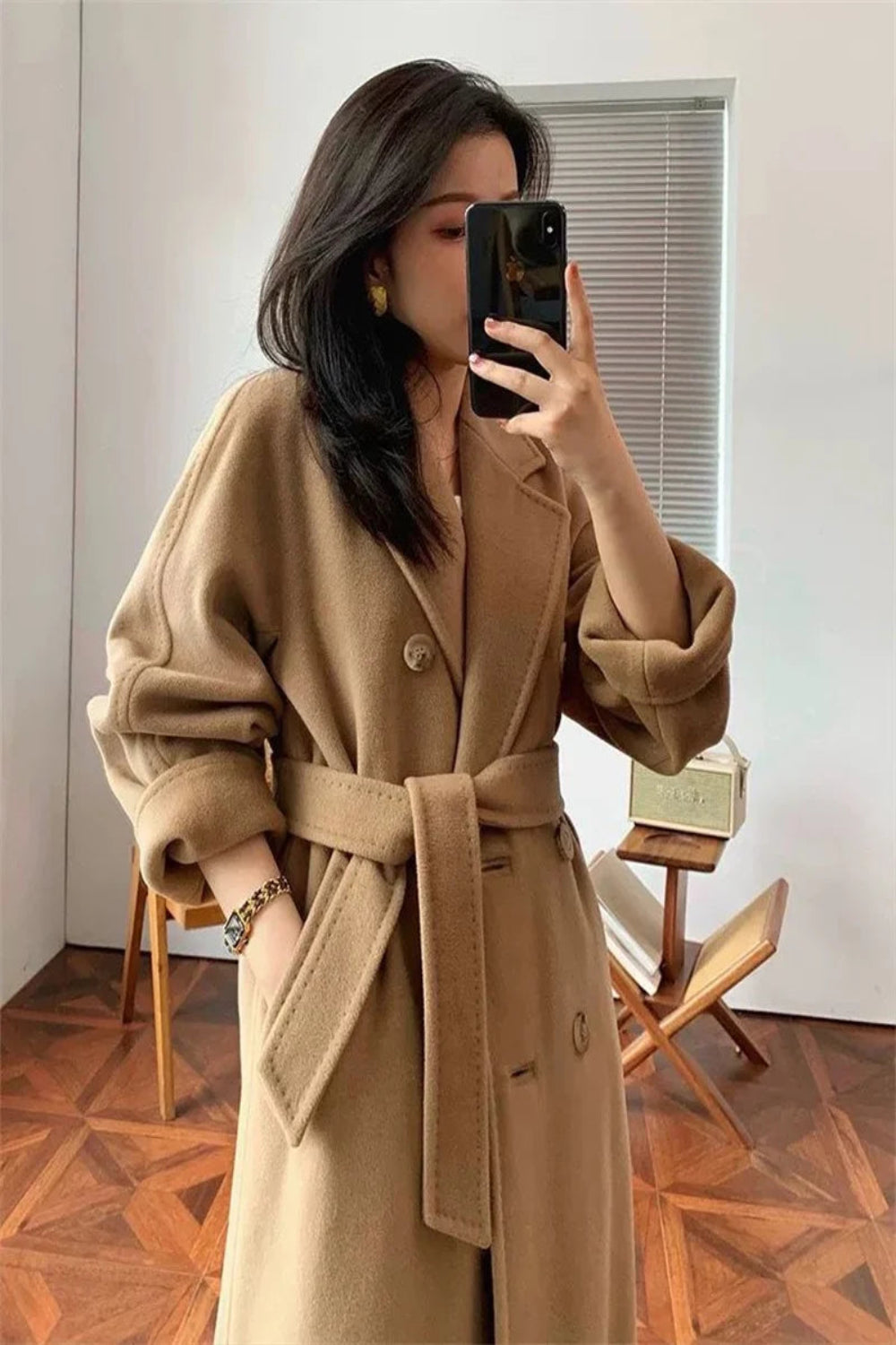 Person wearing a beige trench coat taking a mirror selfie in a room with wooden flooring.