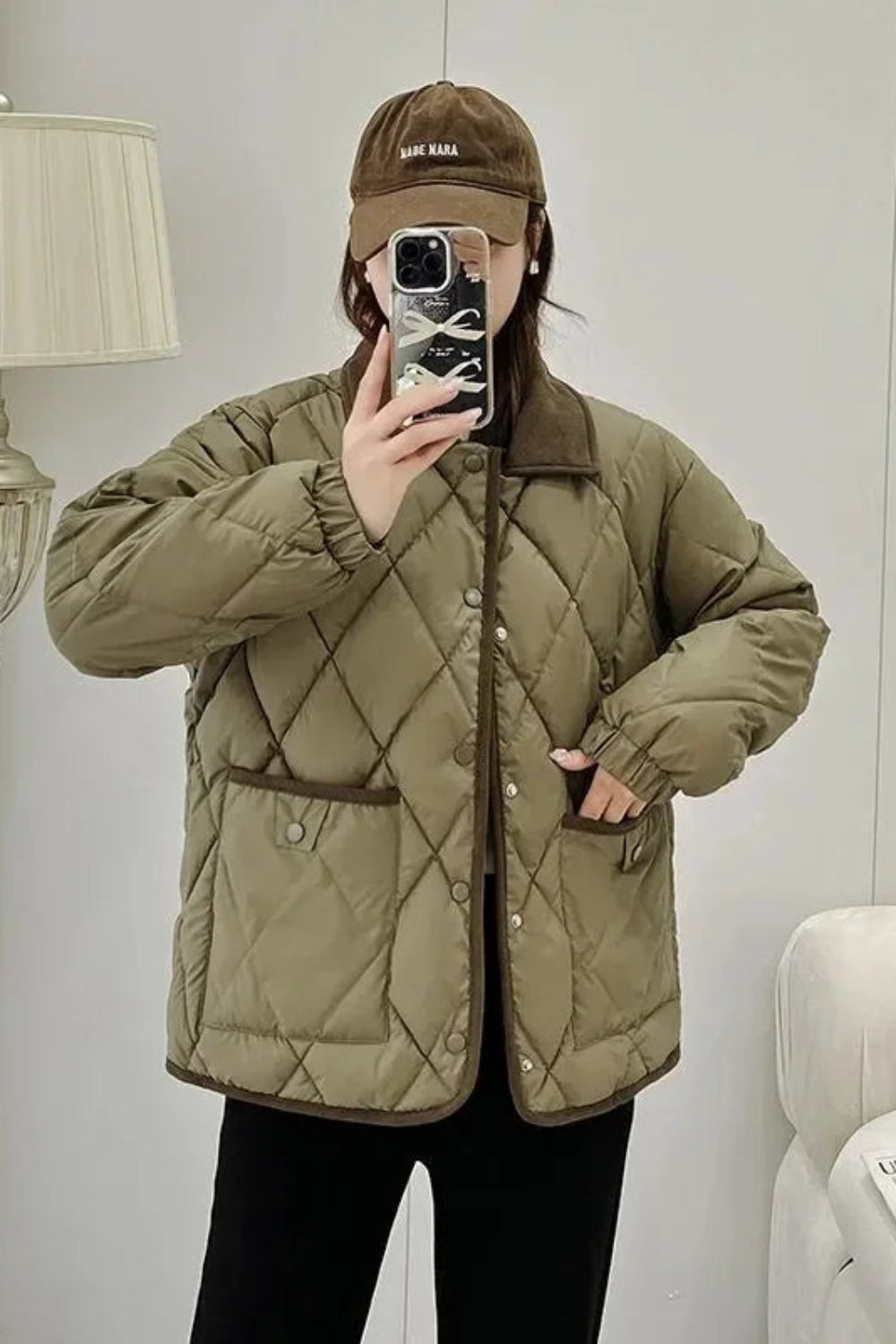 Person wearing a quilted green jacket and brown cap taking a mirror selfie.