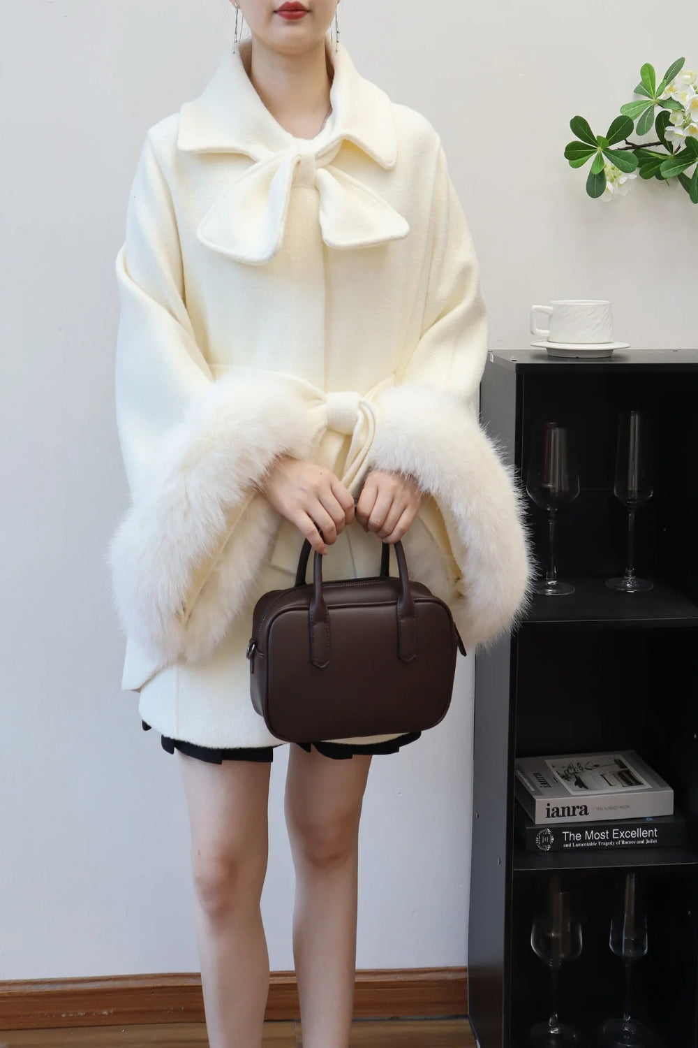 Person wearing a white coat with a large bow, holding a brown handbag indoors.