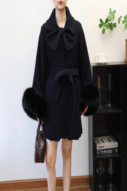 Person wearing a black coat with fur trim in a room with a bookshelf and plant.