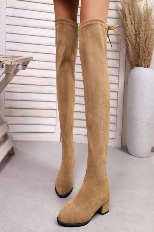 Tan knee-high boots worn by a person on a light gray floor with a white wall and decorative plant in the background.