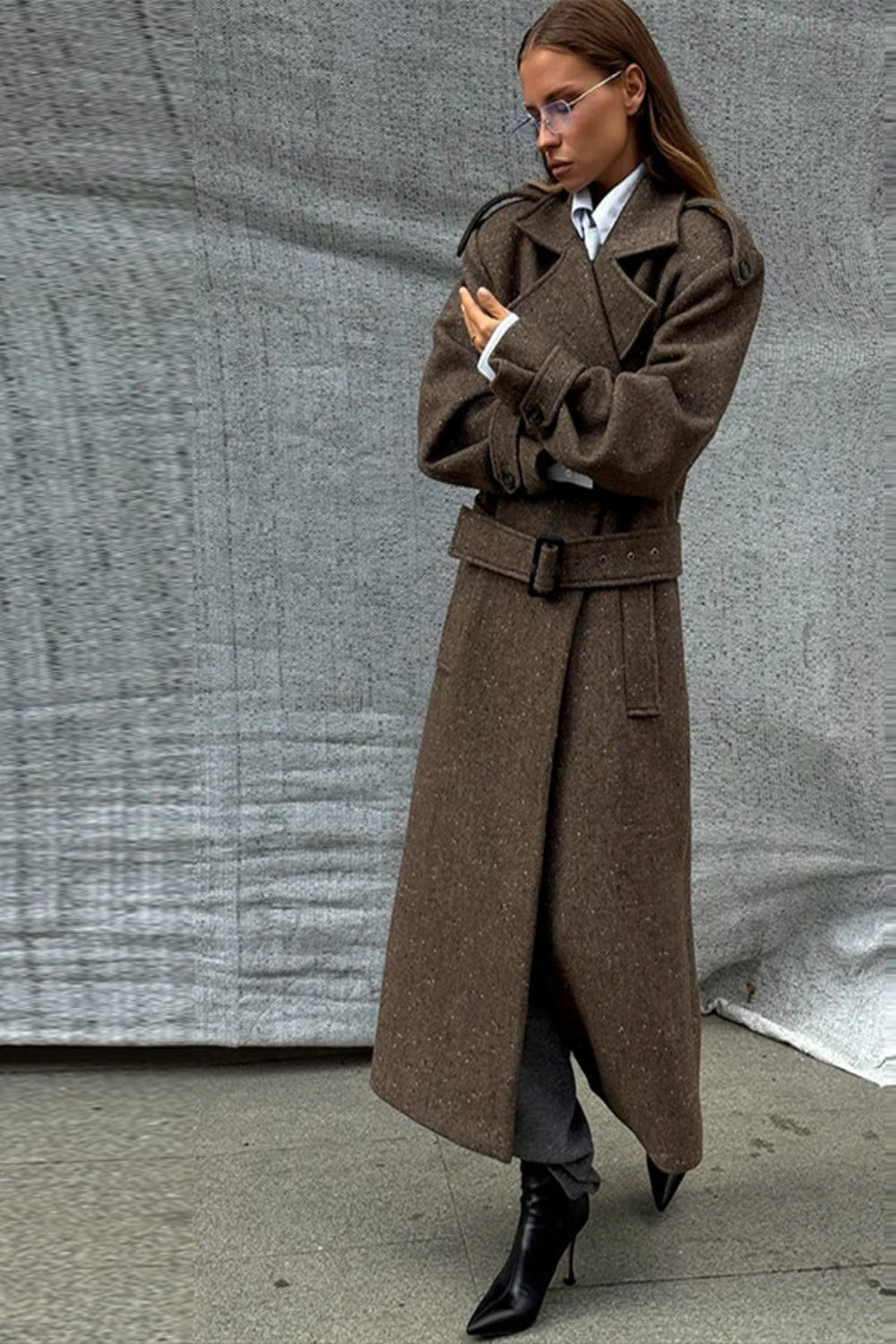 Woman wearing a long brown coat against a gray textured wall.