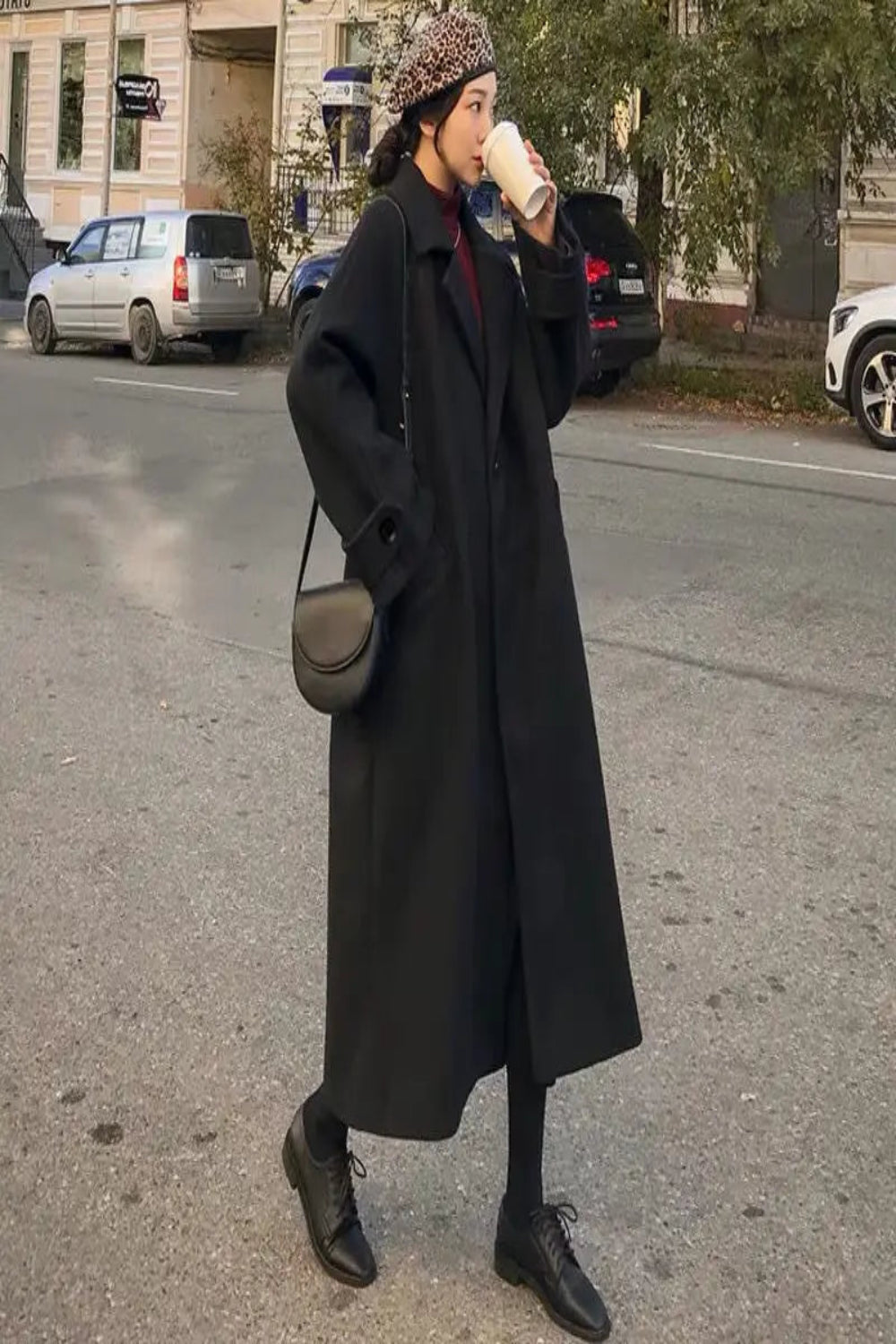 Person wearing a long black coat and patterned beanie drinking from a cup on a street.
