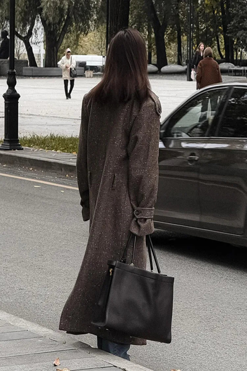 Person walking on a street holding a black bag, wearing a long coat.