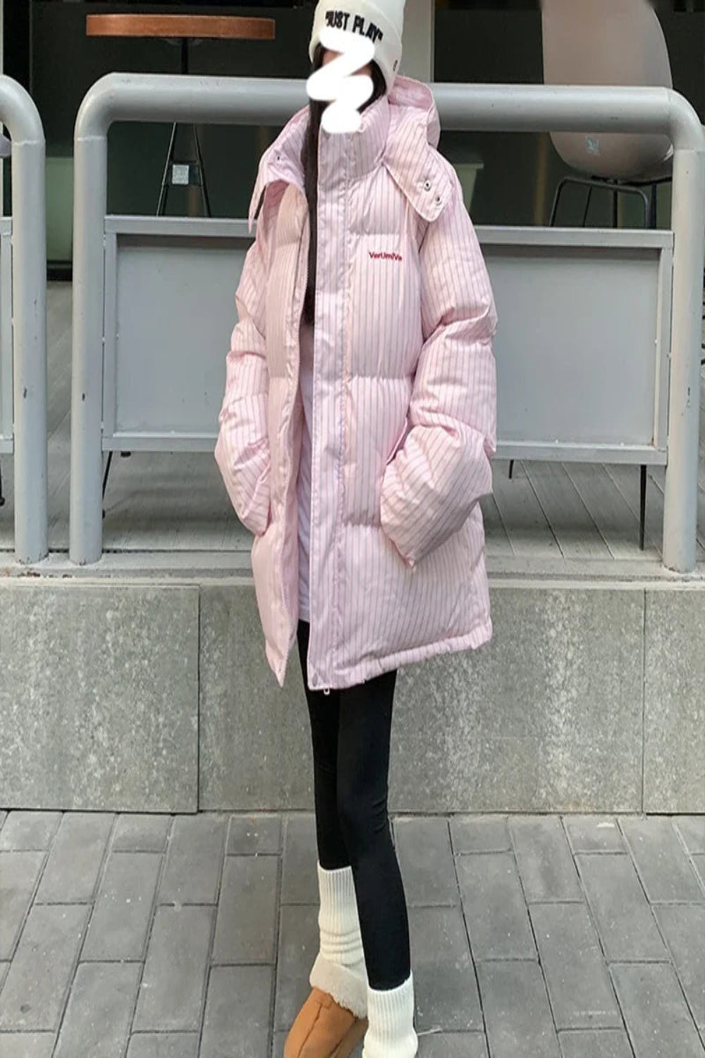 Person wearing a pink puffer jacket with a brand logo, standing on a sidewalk.