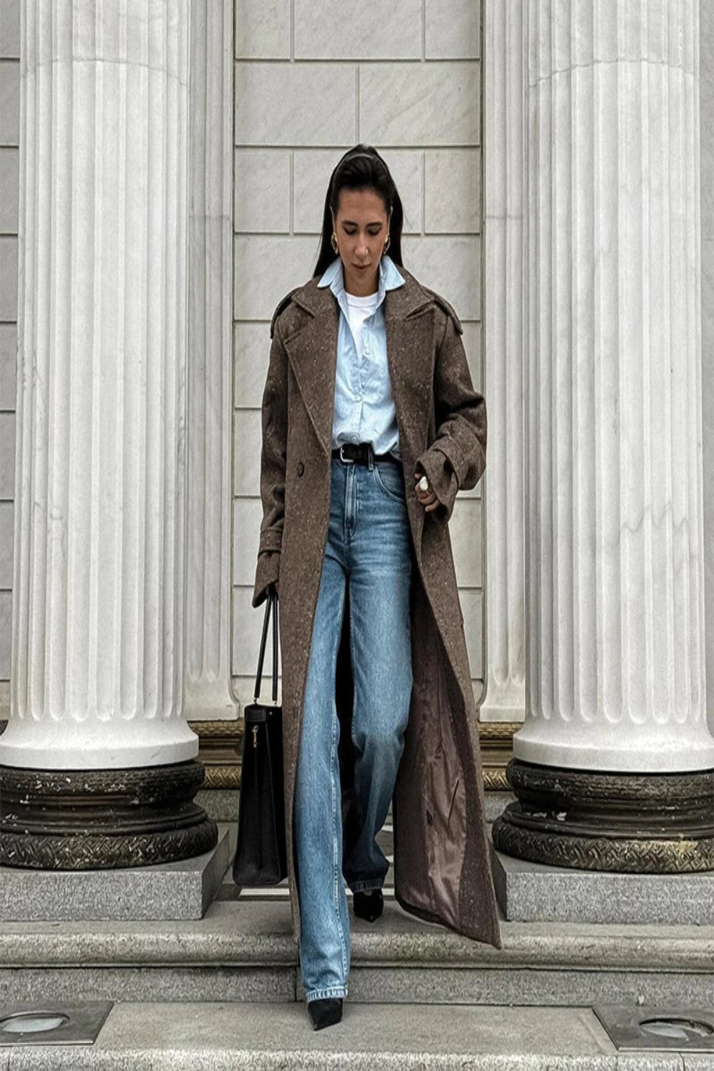 Woman in a brown coat and blue jeans standing on steps with classical columns in the background
