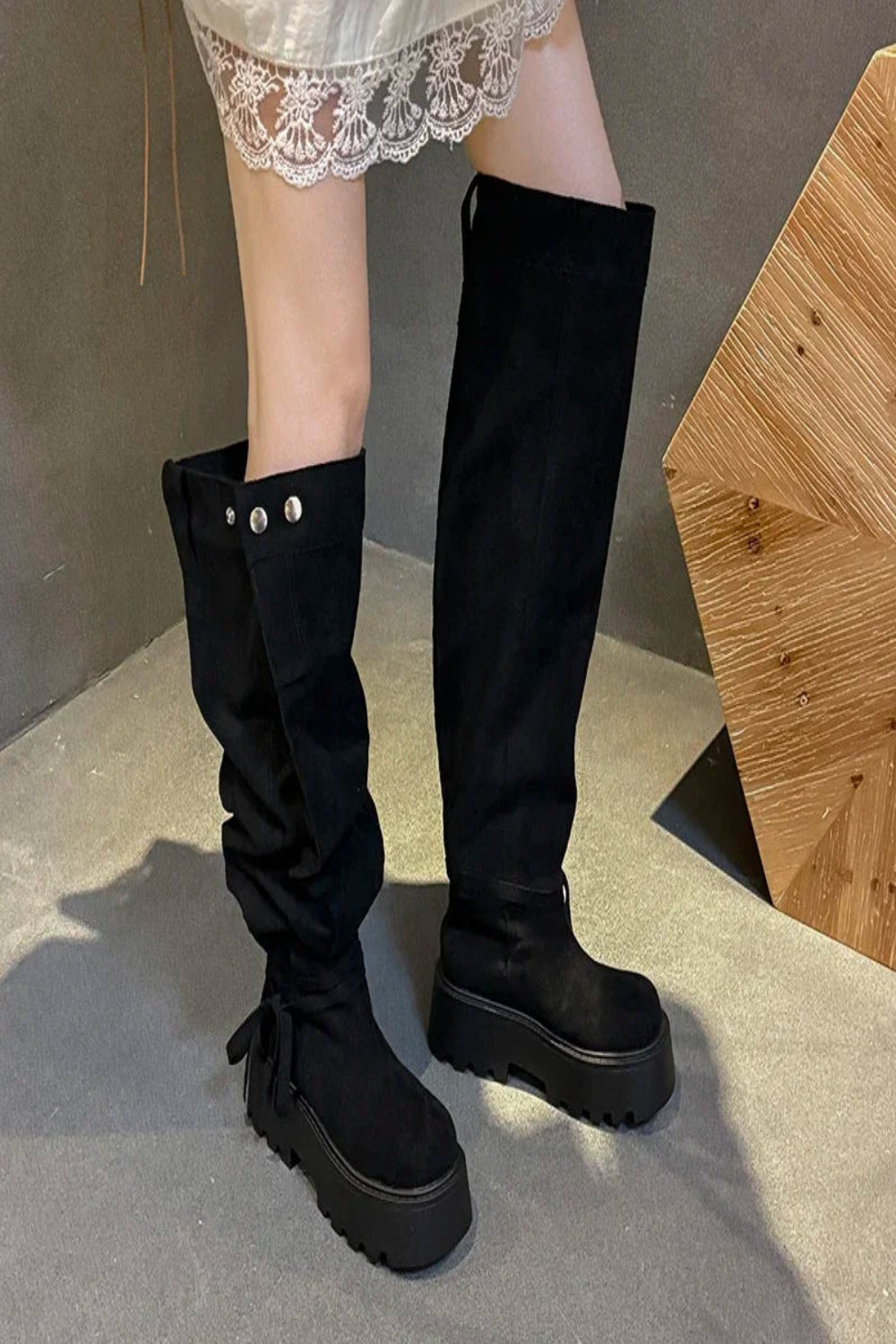 Black knee-high boots worn with a lace skirt on a neutral background