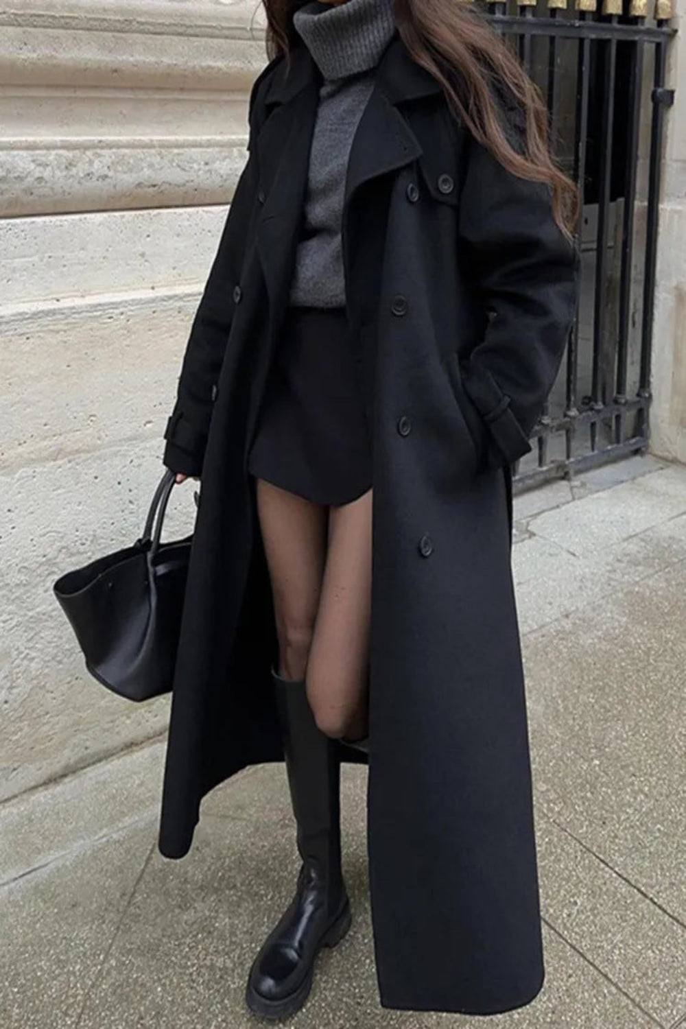 Person wearing a long black coat with a black handbag on a city street.