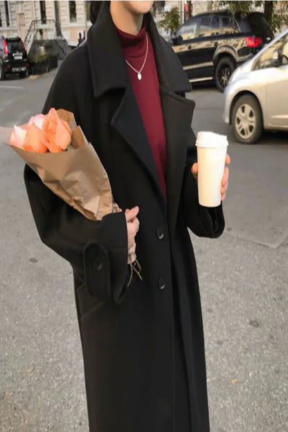 Person in a black coat holding flowers and a coffee cup on a street.