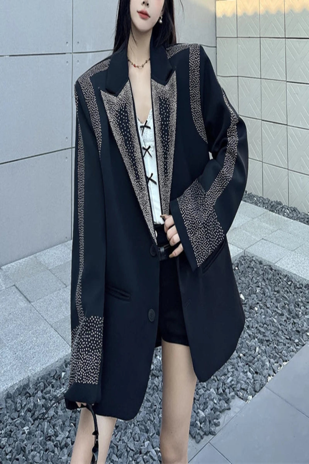 Person wearing a black blazer with decorative patterns standing against a tiled wall.