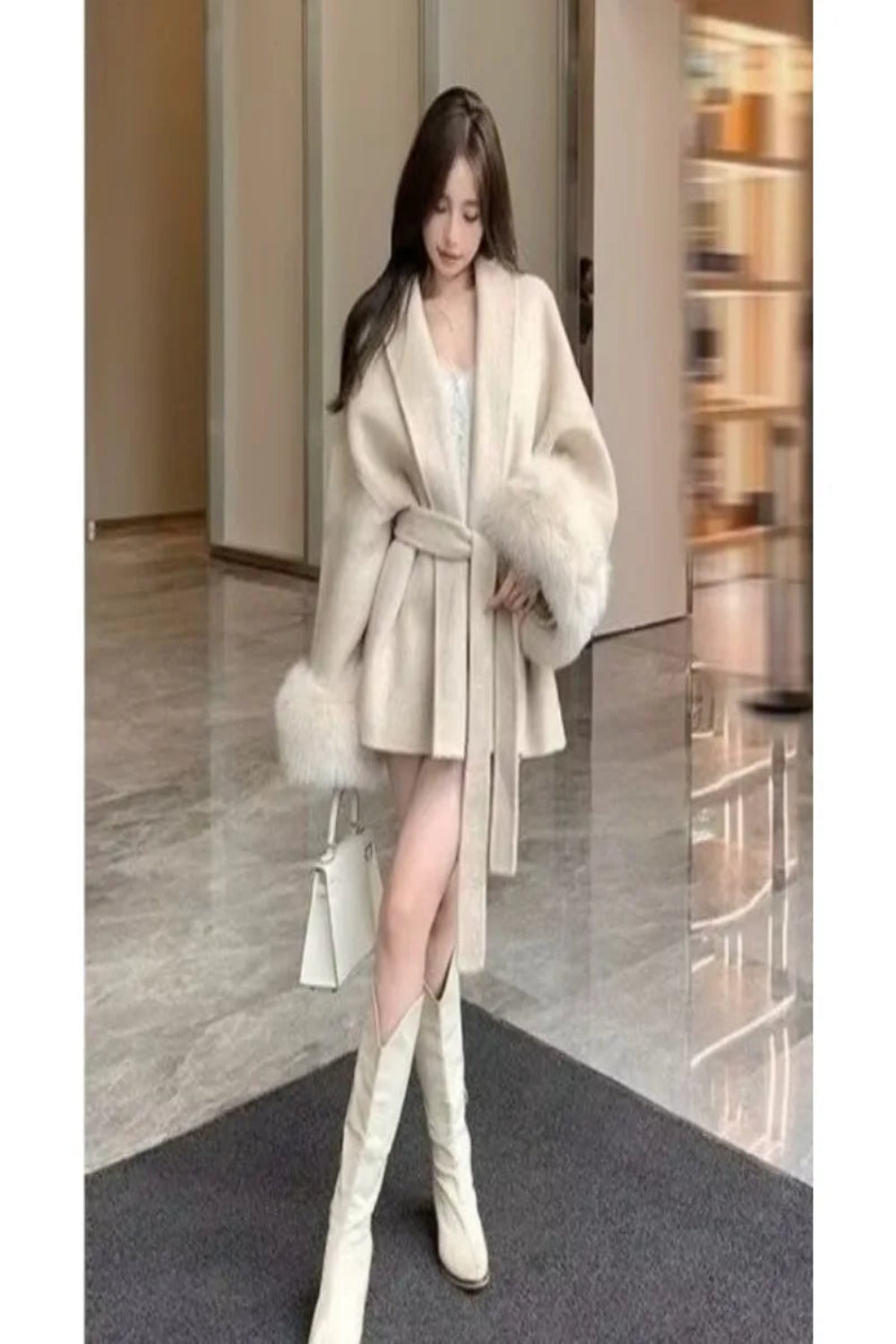 Woman wearing a cream coat with fur trim and white knee-high boots in an indoor setting.