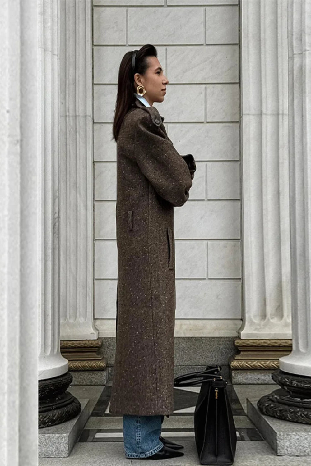 Woman in a long brown coat standing in front of classical columns.