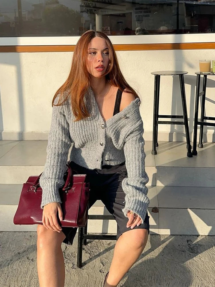 Woman in a gray cardigan holding a red handbag on a beach.