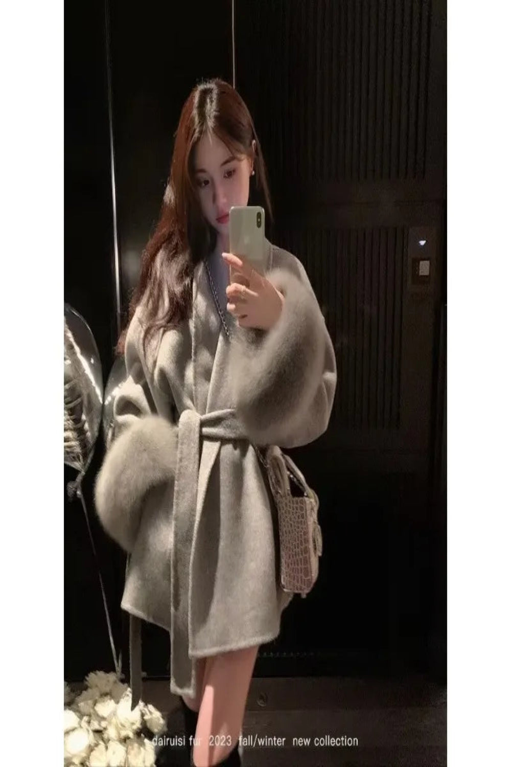 Person wearing a light gray coat taking a mirror selfie in a dimly lit room.