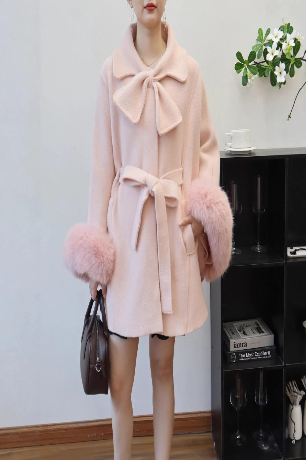 Person wearing a pink coat with fur cuffs and collar, holding a brown handbag indoors.