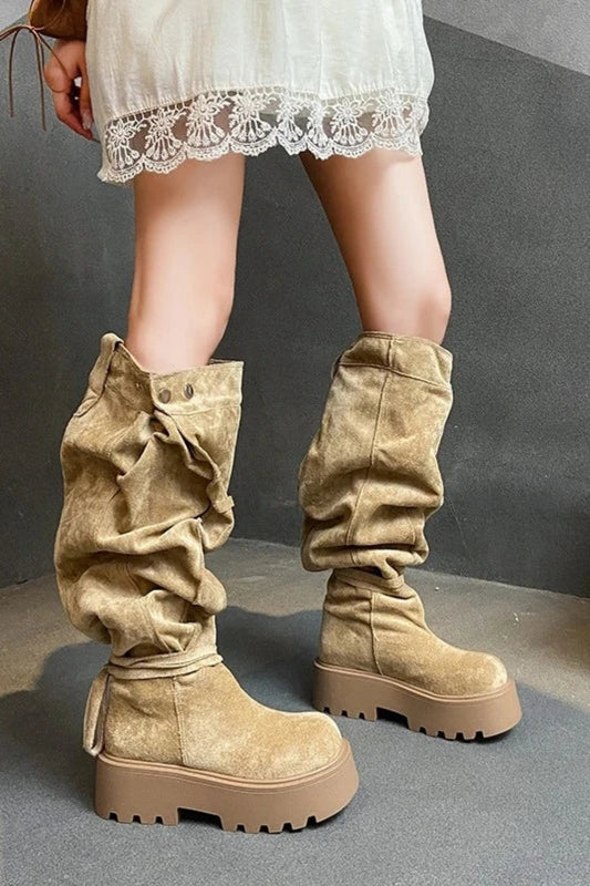 Beige knee-high boots worn with a white lace-trimmed dress on a gray floor.