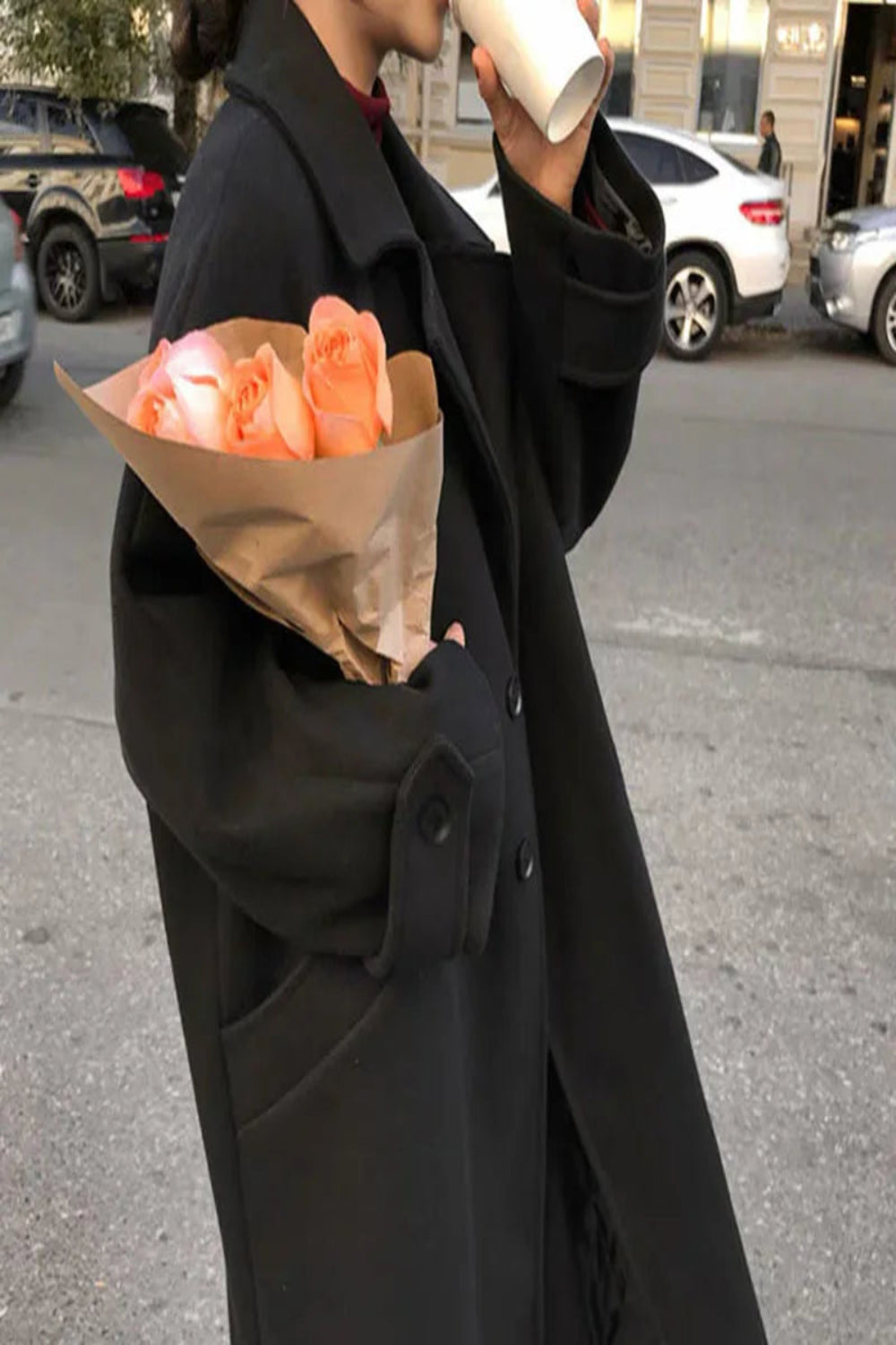 Person in a black coat holding a bouquet of flowers and a cup on a city street.