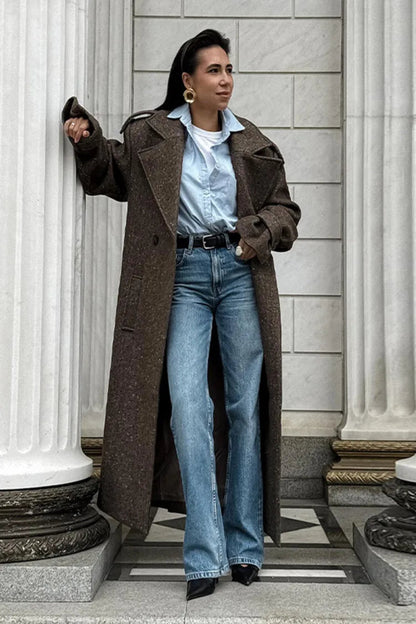 Woman wearing a long brown coat, light blue shirt, and jeans standing in front of classical architecture.