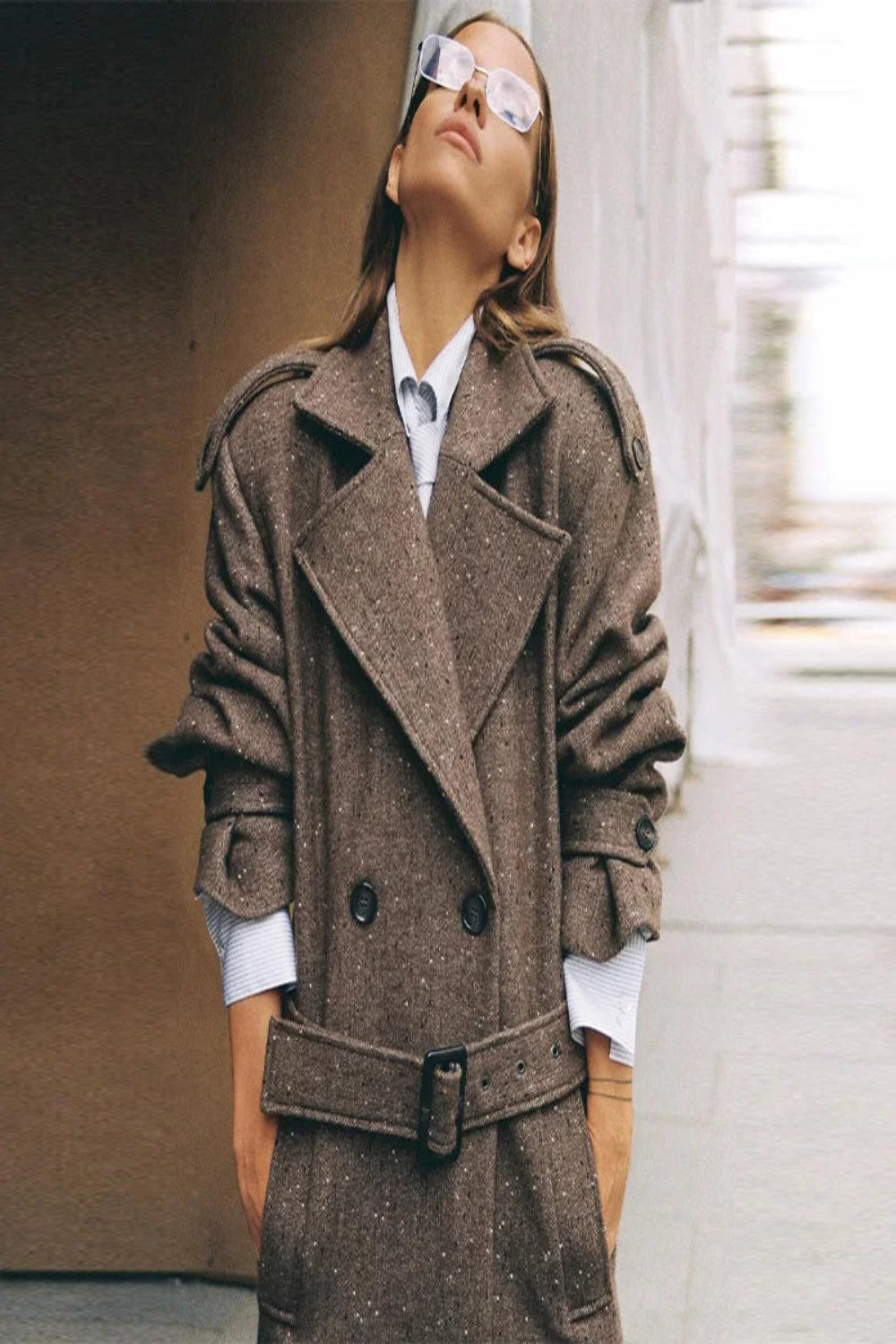 Person wearing a brown coat and sunglasses, standing against a blurred background