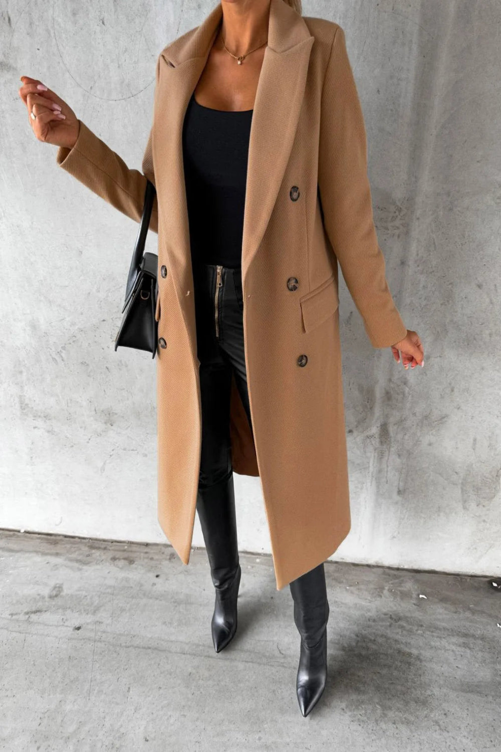 Person wearing a long beige coat, black top, and black boots against a gray concrete wall.