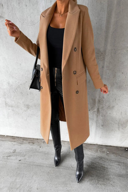 Person wearing a long beige coat, black top, and black boots against a gray concrete wall.