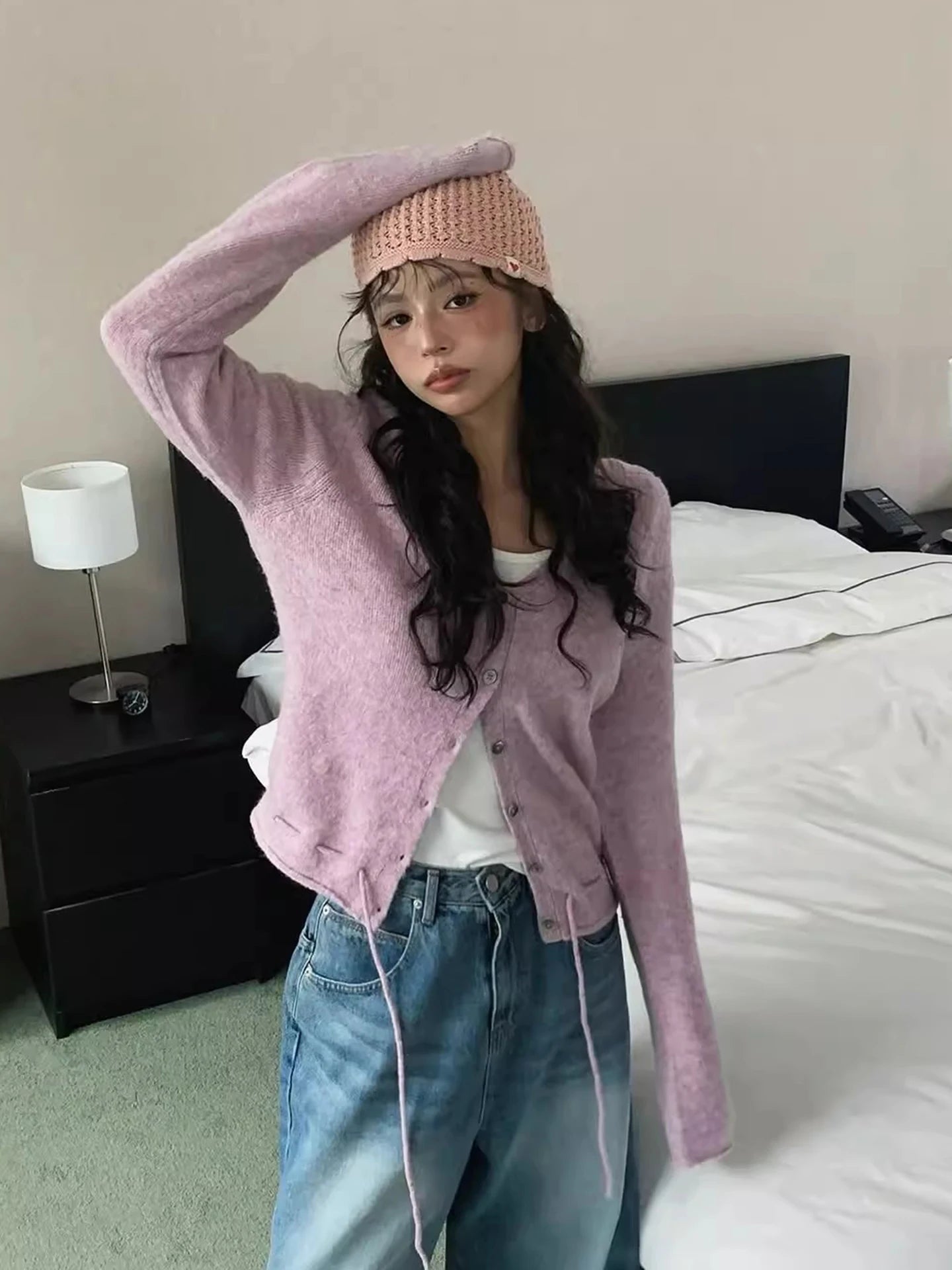 Person wearing a pink cardigan, white shirt, and blue jeans in a bedroom setting.