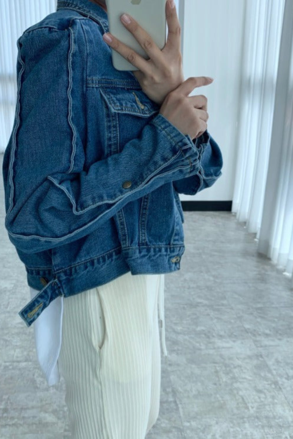 Person wearing a blue denim jacket taking a mirror selfie in a minimalistic room.
