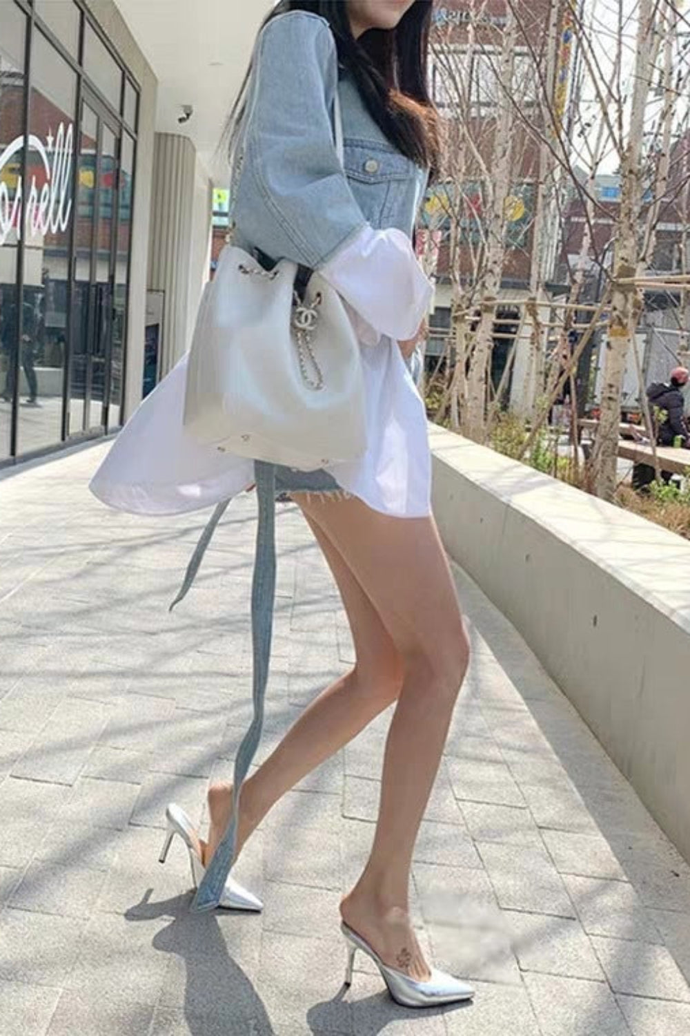 Woman walking outdoors holding a white backpack, wearing a denim jacket and white outfit.
