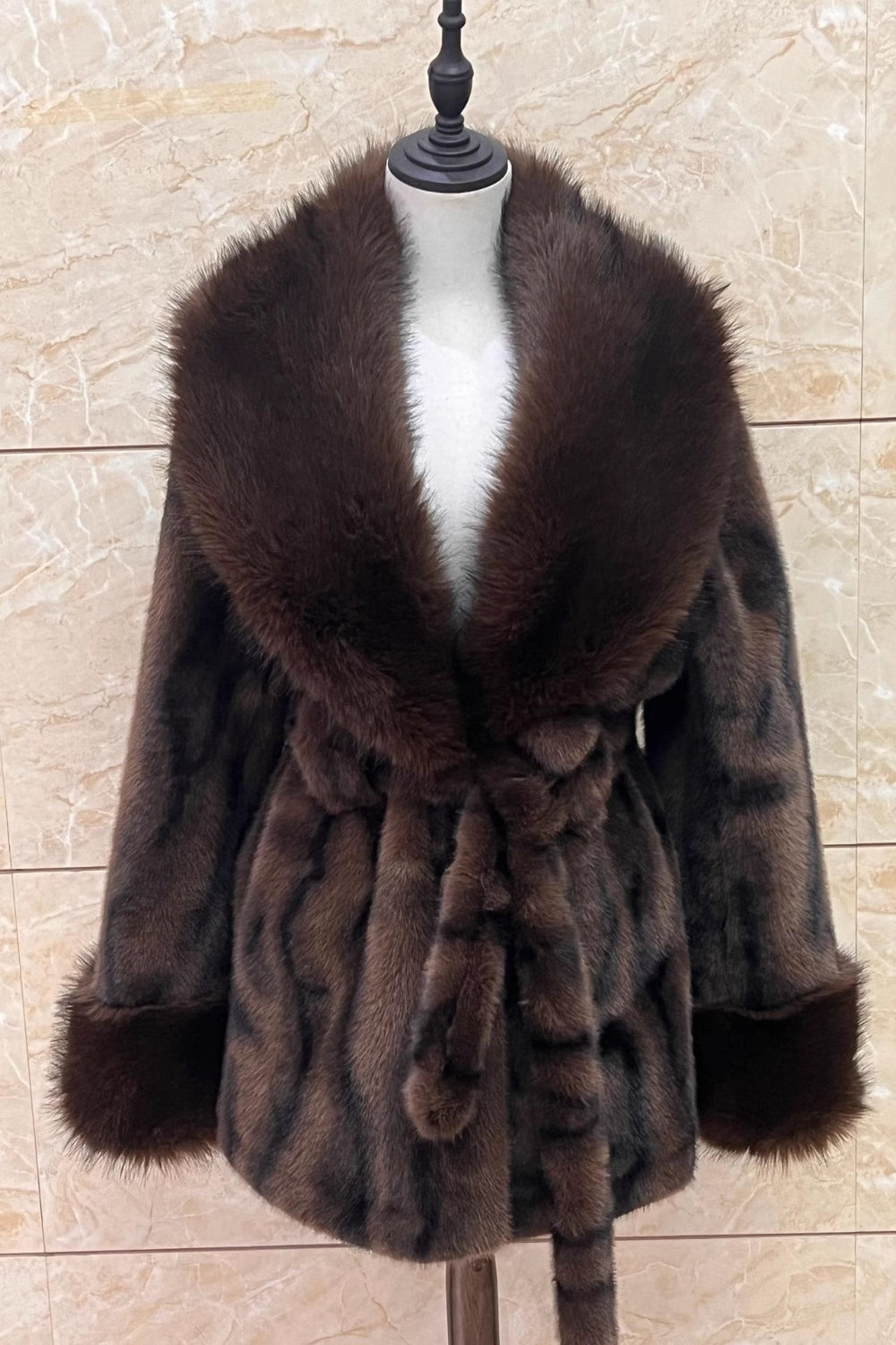 Brown fur coat with a belt on a mannequin against a beige wall.