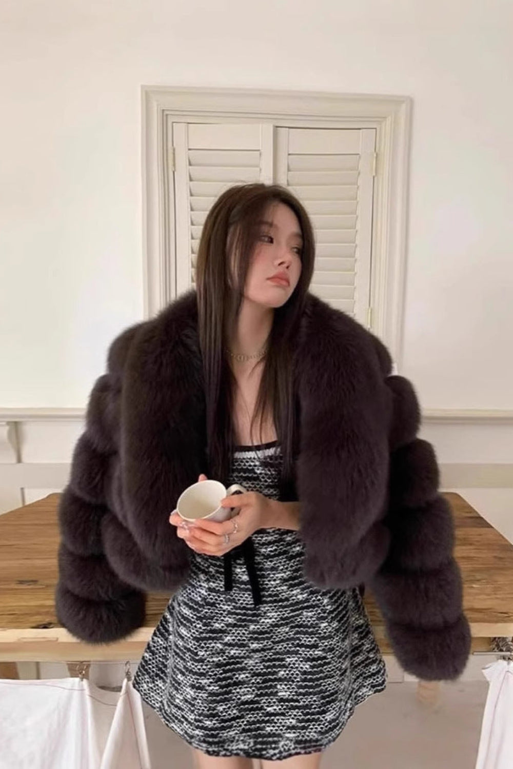 Woman wearing a fur coat in a room with a wooden table and white walls.