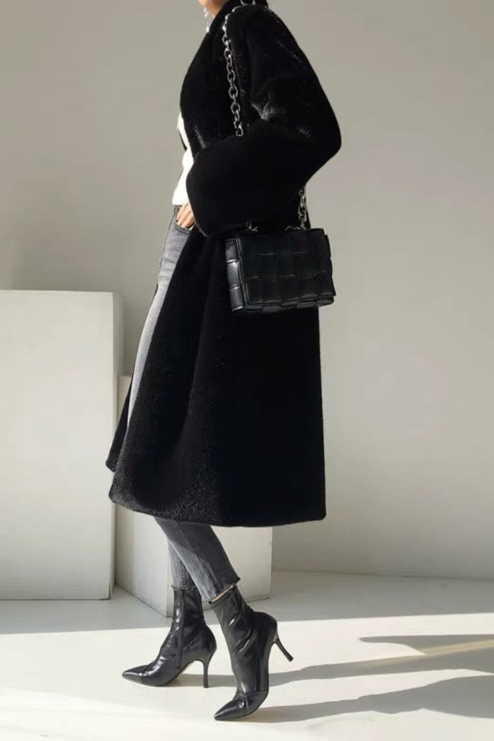 Person wearing a black coat and holding a black handbag in a minimalistic setting
