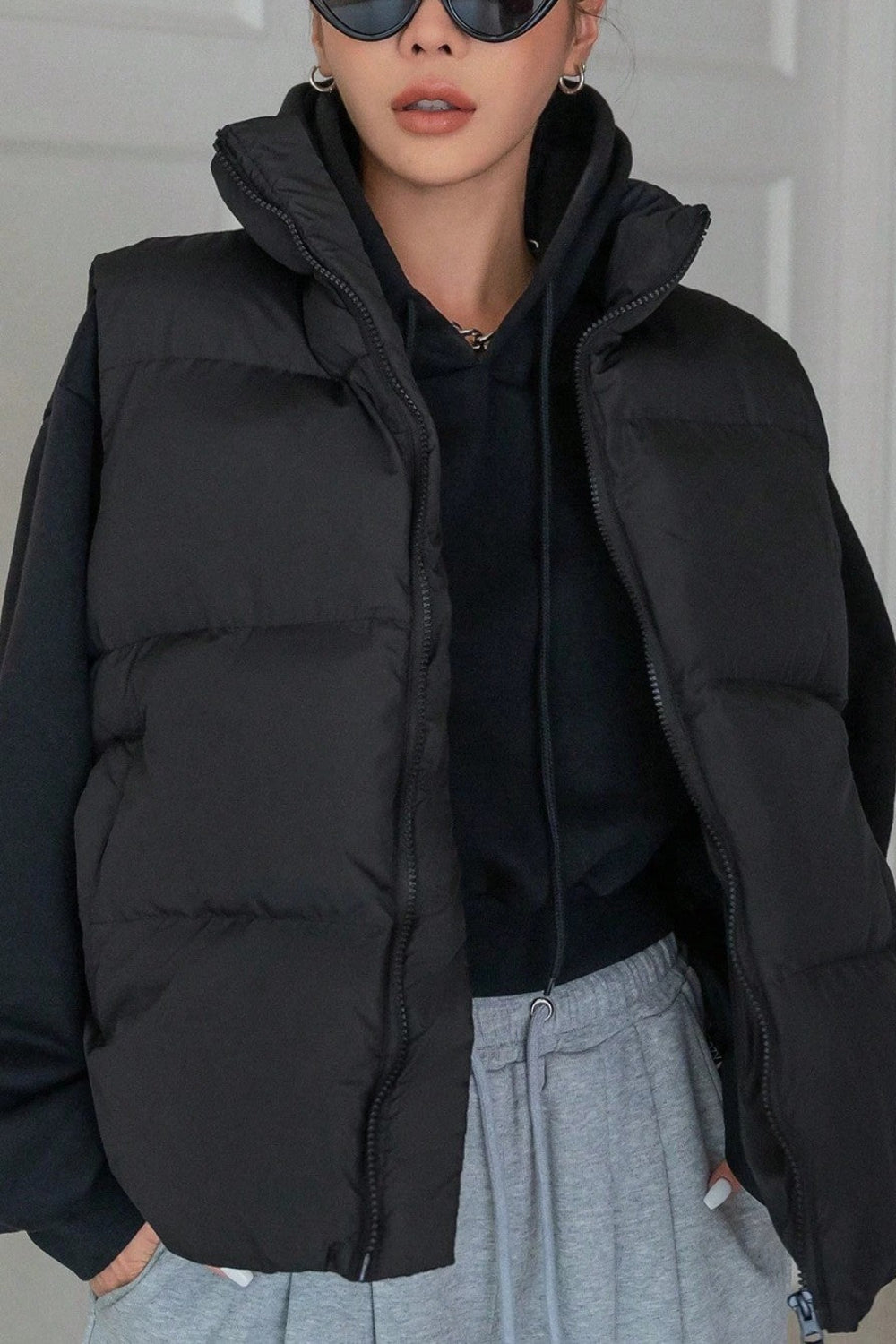 Person wearing a black puffer vest over a black hoodie with gray pants against a neutral background