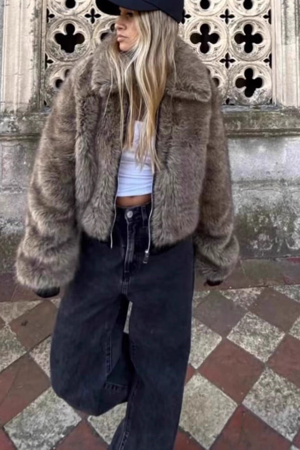 Person wearing a fur coat standing in front of a textured wall with decorative elements.