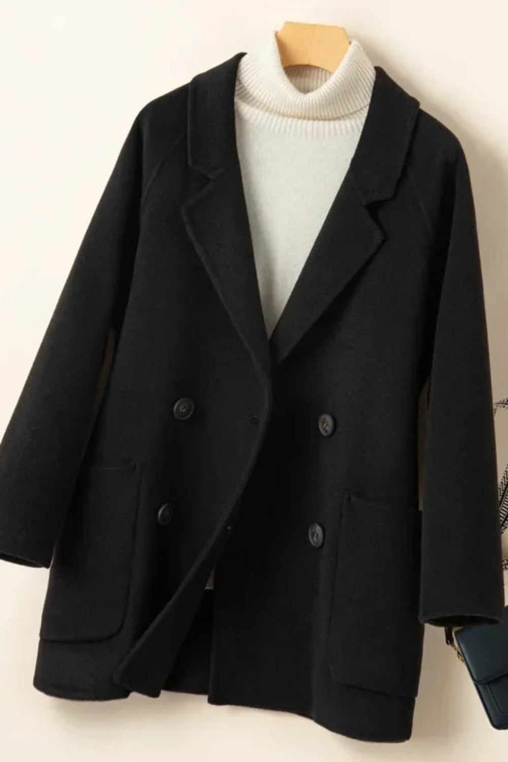 Black coat on a wooden hanger with a white sweater underneath, on a light beige background.