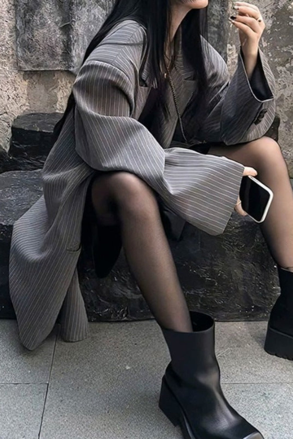 Person wearing a gray coat and black ankle boots sitting on a stone bench.