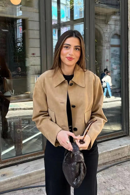 Woman in a beige coat holding a black bag outdoors near a building entrance.