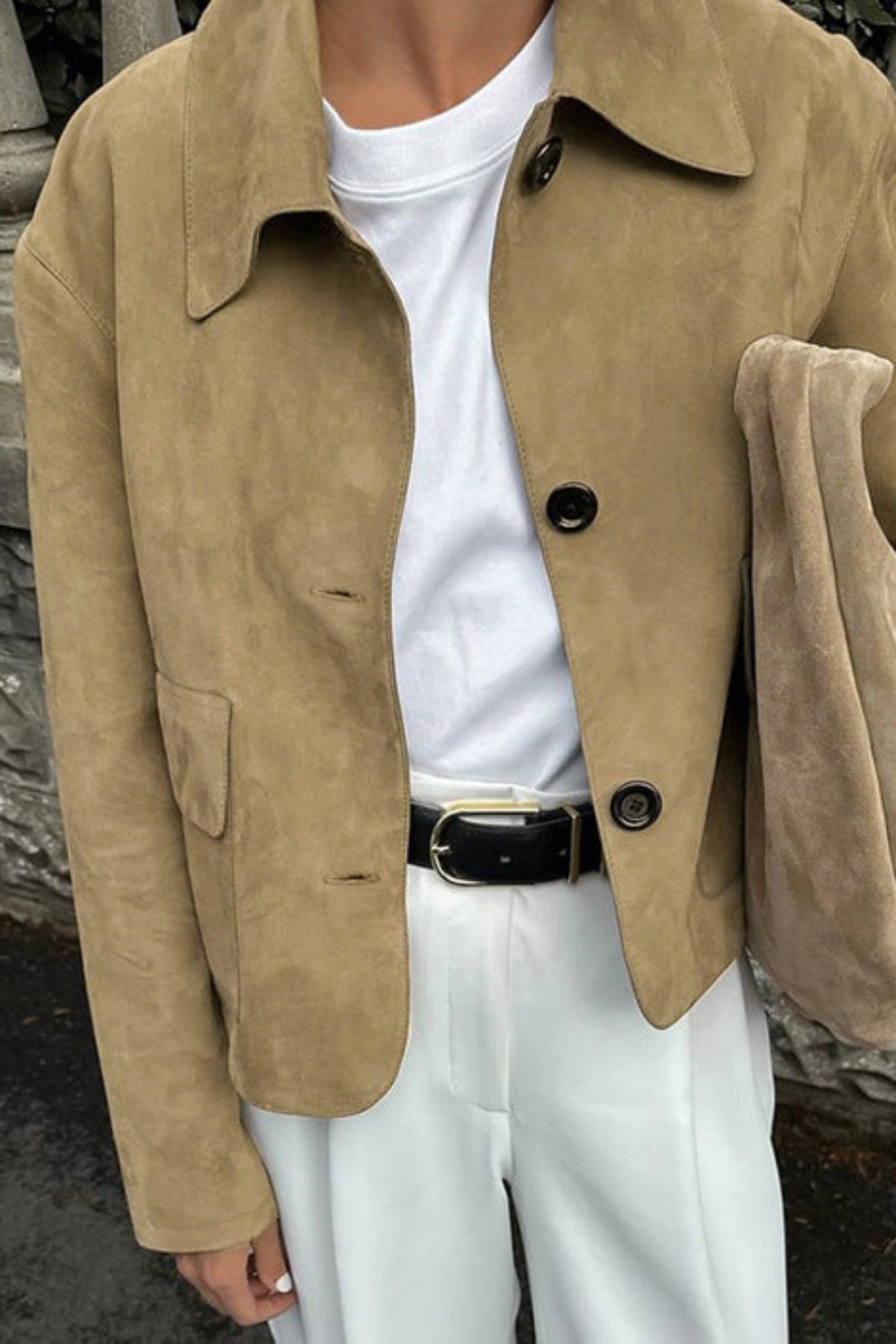Person wearing a beige jacket over a white shirt and pants, standing against a stone wall.