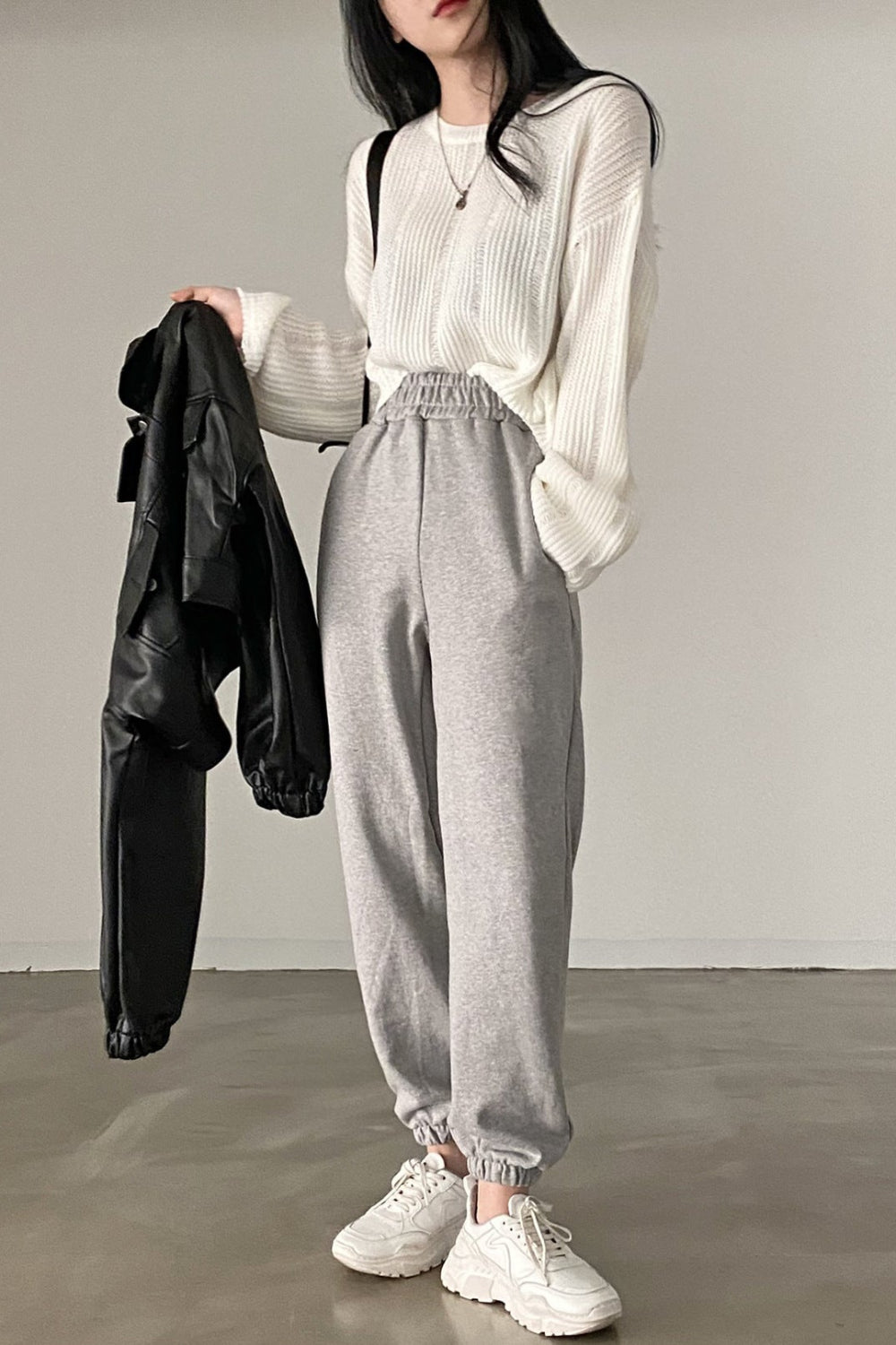 Person wearing a white knit sweater, gray high-waisted pants, and white sneakers, holding a black jacket.