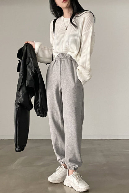 Person wearing a white knit sweater, gray high-waisted pants, and white sneakers, holding a black jacket.