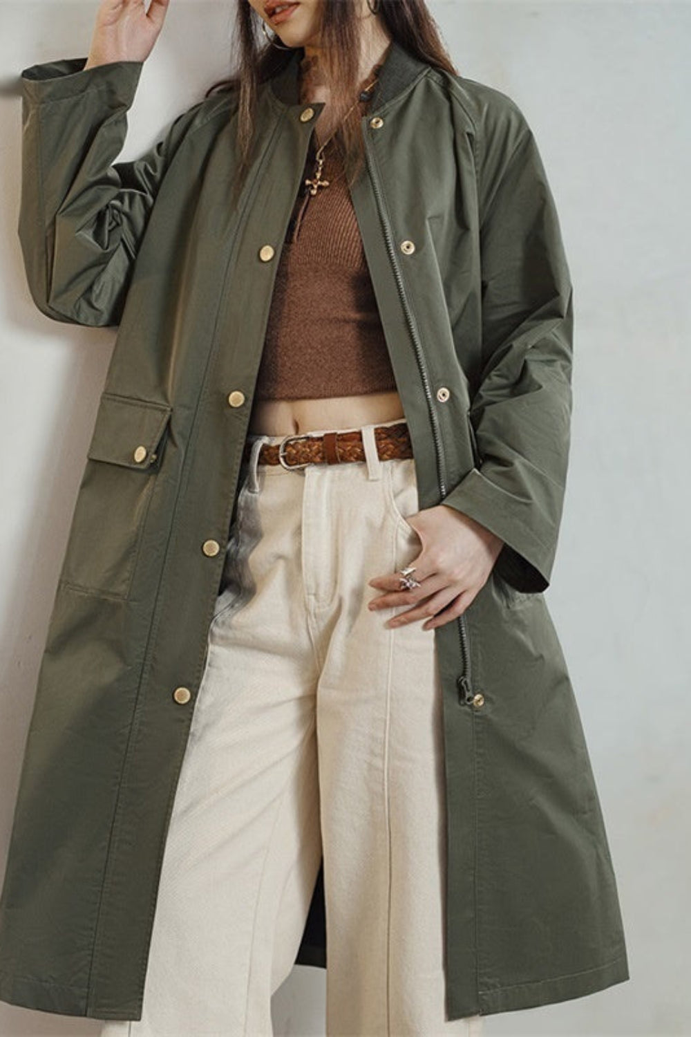 Person wearing a green trench coat over a brown sweater and white pants on a plain background
